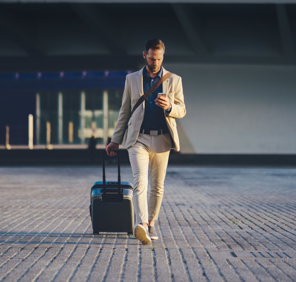 A man in a beige suit walks with a wheeled suitcase while looking at his phone.